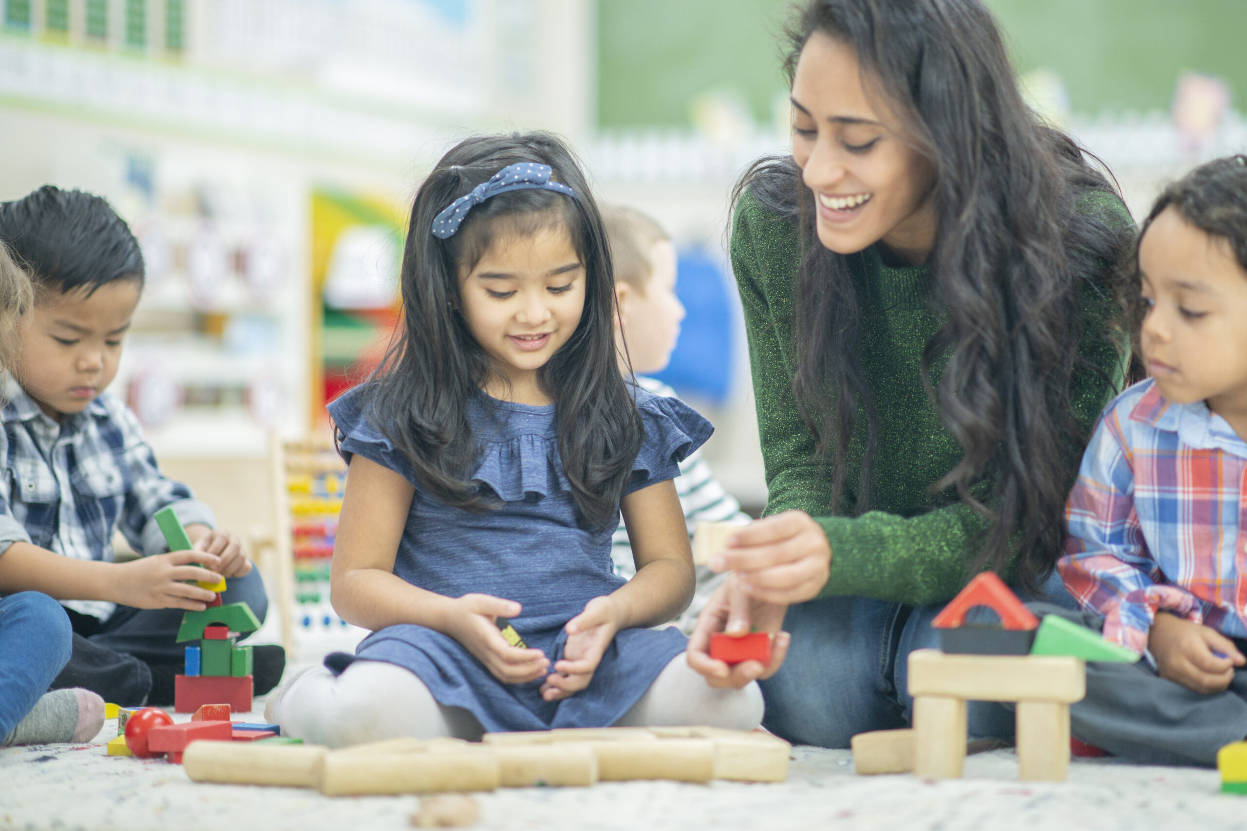 A young beautiful teacher is  playing with her students. She has a giant smile on her face while she holds a block in her hand.